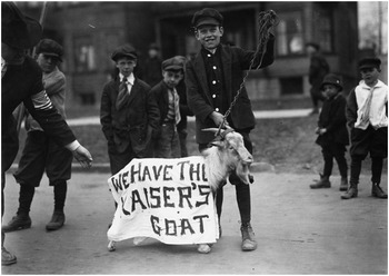 A photo depicts a boy leading a goat in the middle of the street. The goat is draped in a white sheet, and the words, We have the Kaiser's goat, are written on it. Other children are observing the scene. See long description.