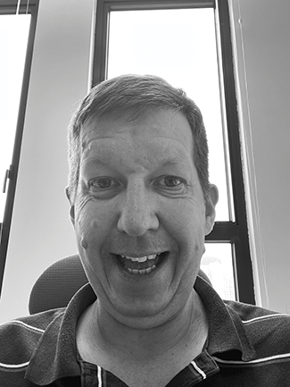 A person with short brown hair, wearing a dark polo shirt, smiles broadly while standing in front of a window.