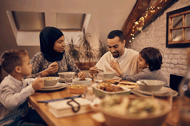 A man, a woman and two children sit around a dining table in a living room. The table is filled with several dishes and the group appears to be sharing and enjoying the meal. The woman wears a headscarf and smiles.