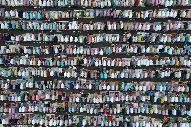 An aerial view showing 11 rows of Muslims in prayer, each row consisting of approximately 40 to 50 individuals. They are in a prostrated position on prayer mats, wearing long garments that cover their bodies and heads.