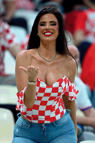 A young woman with long dark hair smiles at the camera while leaning forward in a sports stadium. She is wearing a check-print sweetheart neckline top and a gold necklace. She appears to be part of the audience.