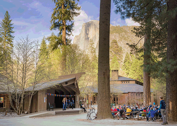 Performers captured mid-performance on an outdoor stage, with benches arranged among trees and the granite Half Dome visible in the background. Part of the Shakespeare in Yosemite setting in Curry Village, Yosemite National Park.