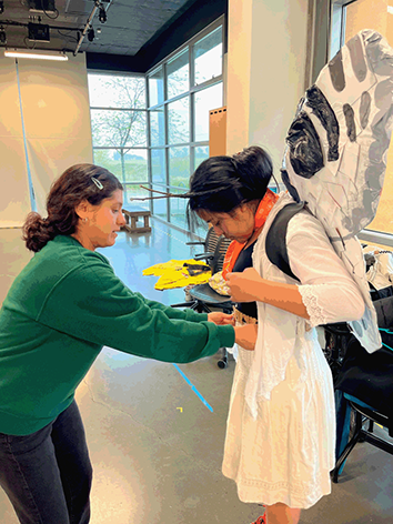 A young woman in a sweatshirt assists another woman, who is wearing a dress and fairy wings made of trash, in fitting a belt.