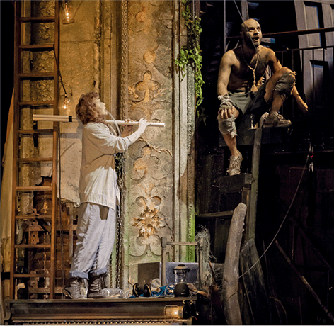 A woman playing an instrument made from discarded plastic tubing, while a shirtless man portraying Caliban sings.
