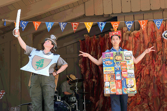 A park ranger in uniform and knight’s armor made of cardboard holds up a fake sword, while a nonbinary actor, dressed in a fake wall made of cereal and cracker boxes, holds up two fingers with both hands.