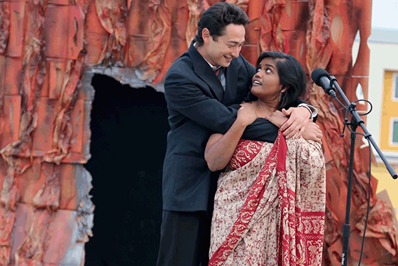 A man in a suit places his arm around a woman in a sari as they stand in front of the base of a sequoia tree made from cardboard and discarded wood.