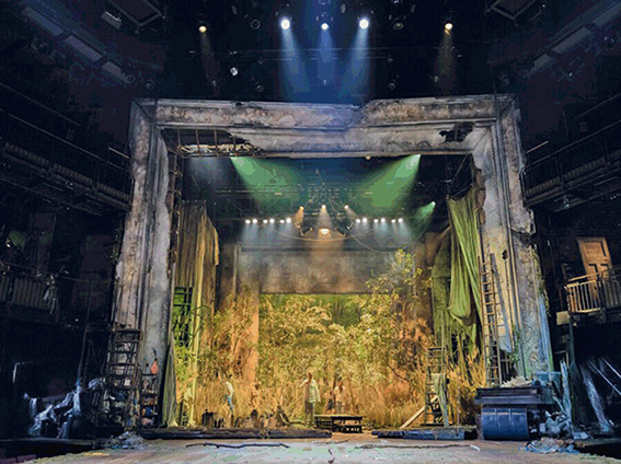 Barely visible actors are surrounded by trees on stage, bathed in golden light, in front of the proscenium arch at the Royal Shakespeare Theatre.