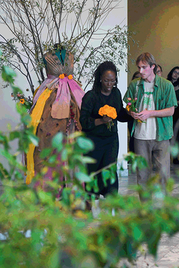 A woman holds flowers while a man looks on, as they stand in front of a dress made of tree branches, flowers, and discarded materials.