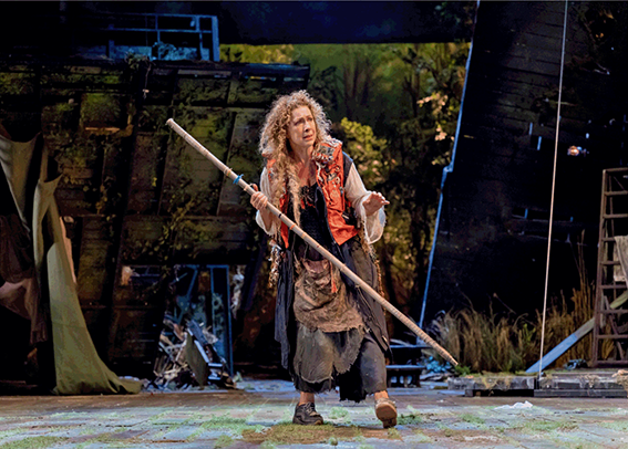 Actor Alex Kingston as Prospero, wearing a life vest and ragged clothing. They are holding a pole or stick, and the background setup suggests vegetation.