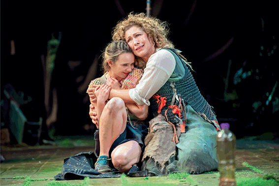 Alex Kingston, portraying Prospero, kneels on the ground beside Jessica Rhodes, who plays Miranda, with her arms around Jessica.