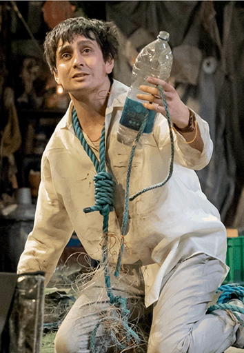 A male actor portraying Ferdinand kneels on stage while holding a plastic water bottle. He has a loose noose tied around his neck.