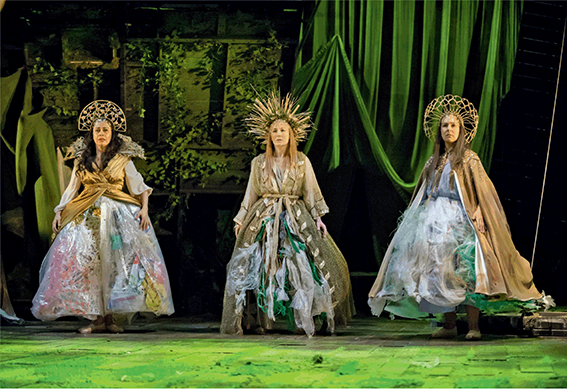 Three women, dressed in dresses made of trash, representing Sky, Earth, and Rain.