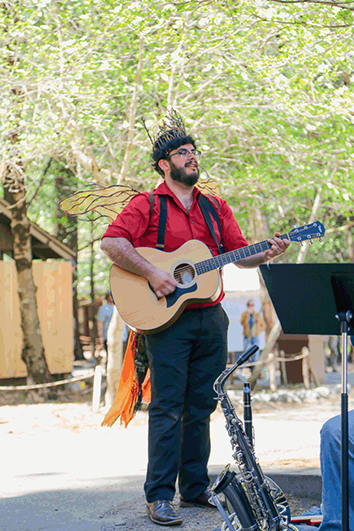 A young bearded man plays guitar, dressed in a shirt, fairy wings, and a crown resembling fire, all made of trash. Several pine trees are visible in the background.