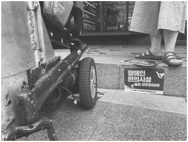 A photograph shows a portion of a wheelchair on the left side. To the right, a sign with Korean text is placed on a step near a person's feet wearing sandals. The background shows a storefront.