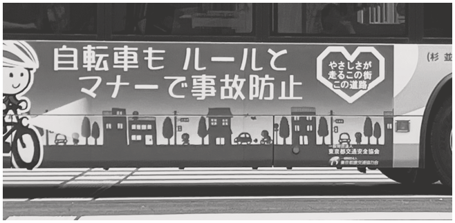 A photograph shows a sign on the side of a bus. The sign includes Japanese text. A cartoon cyclist is on the left. The background of the sign depicts a row of buildings and trees.