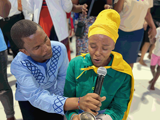 A woman, assisted by a T R S H officer holding a microphone, speaks in the voice of her deceased grandmother during a spiritual service.