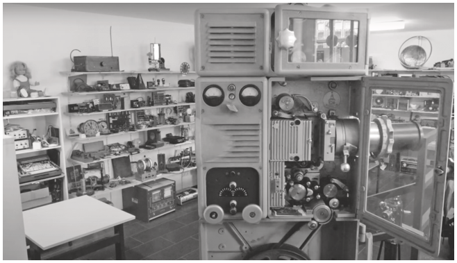 The interior of a room is captured in a photo, showcasing a film projector in the foreground and a collection of mechanical and electronic devices displayed on wooden shelves at the back.