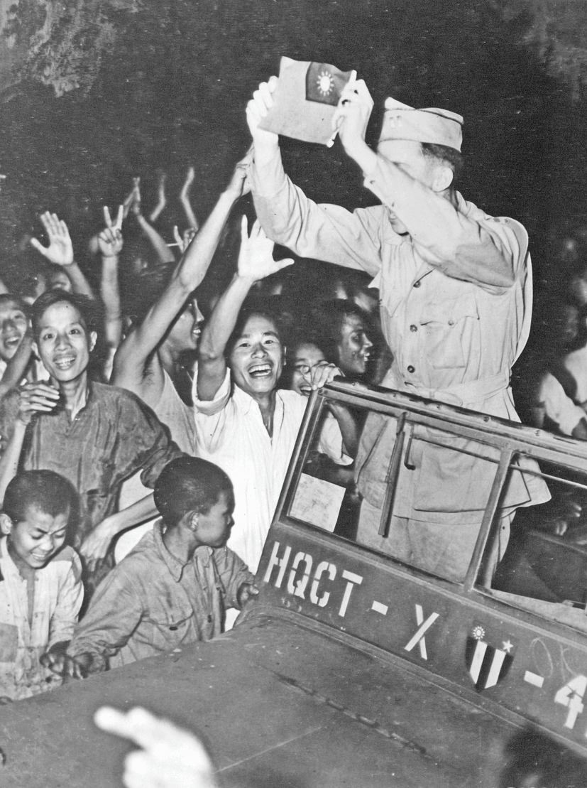 A U.S. Army captain holds a miniature flag of the Republic of China whilst standing on the passenger seat of a military Jeep. He is greeted by cheering Chinese civilians.