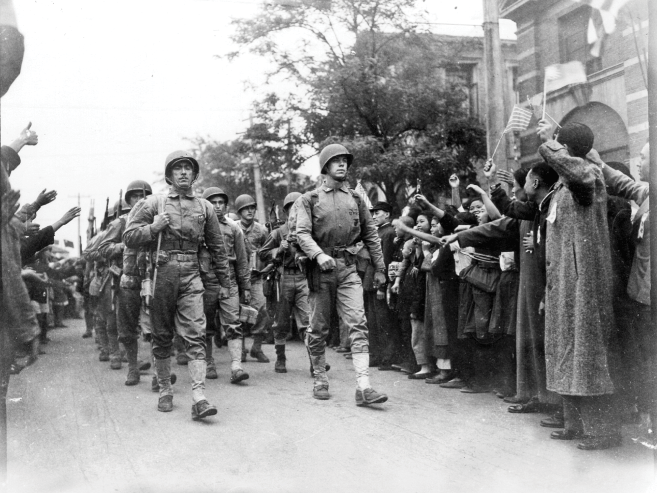 A column of U.S. infantry marches past cheering Chinese civilians, who are waving flags of the United States and Republic of China.