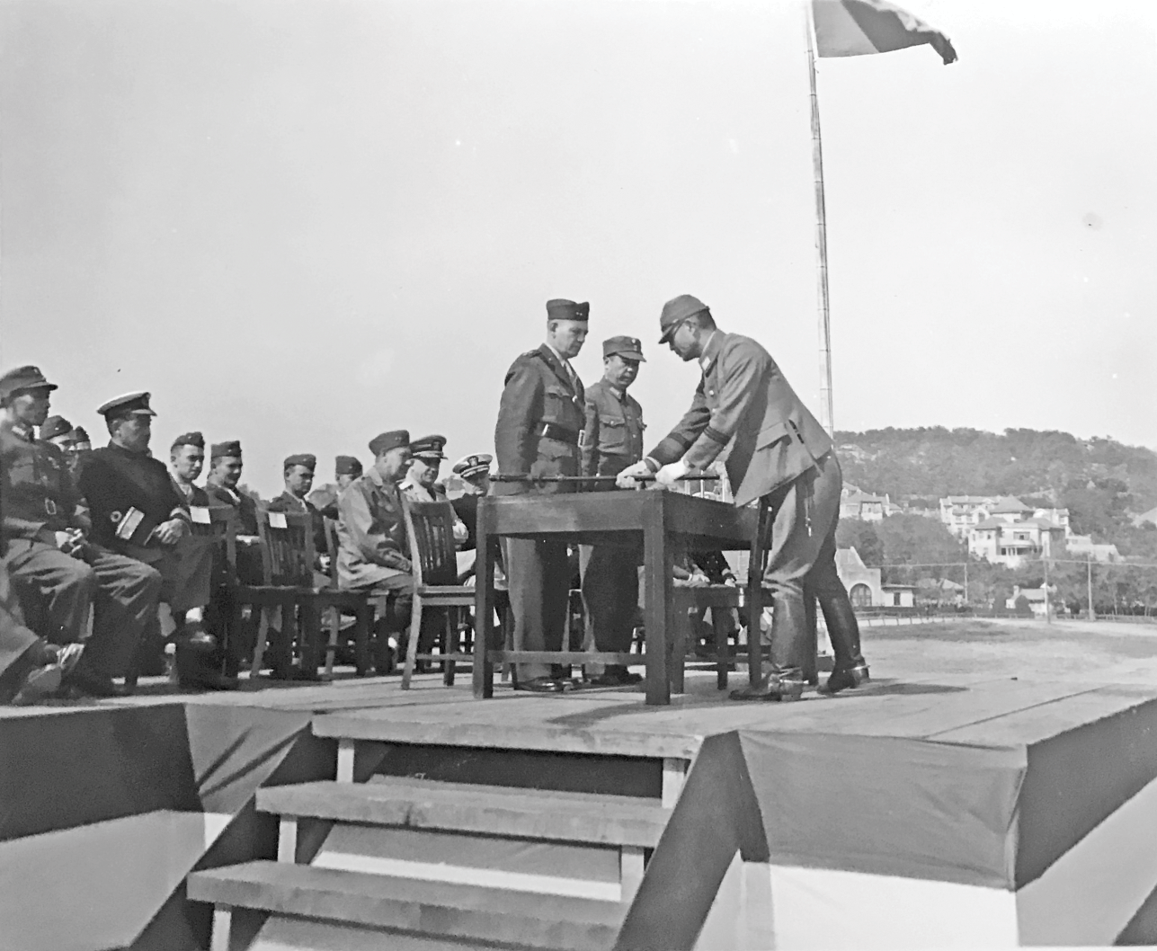 Major General Shepherd and Lieutenant General Chen Baocang stand, accepting the surrender, as the Japanese representative, Major General Nagano Eiji, unbuckles his samurai sword and lays it on the table. See long description.