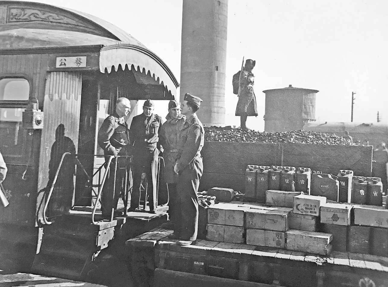 General Dewitt C. Peck, accompanied by two officers, stands on the platform of a railway car, conversing with an American soldier who is on a flat wagon loaded with cargo. See long description.
