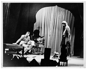 A man sits on a divan holding a script; next to him are an actor in a shopping cart, two mechanical toy monkeys, and a woman in a turban.