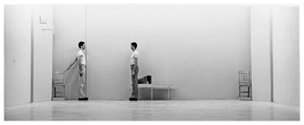 Two men face one another on a wide, nearly empty white stage. Both wear white T-shirts, bell-bottom pants, and shoes with elevated heels. One man extends his arm back toward a wooden chair. The other rests his right leg behind him on a wooden bench.