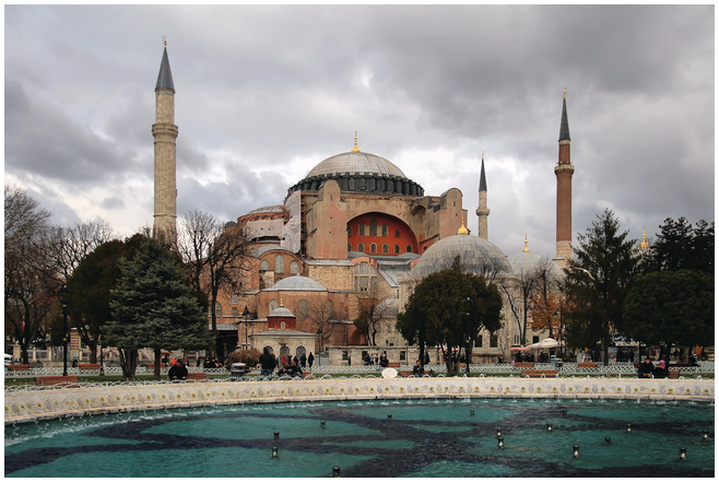A distant view of the domed structure of Hagia Sophia. It stands in the middle field surrounded by trees and 3 vertical minarets. The building is set against a cloudy sky in the background and a wide pool of water in the foreground.