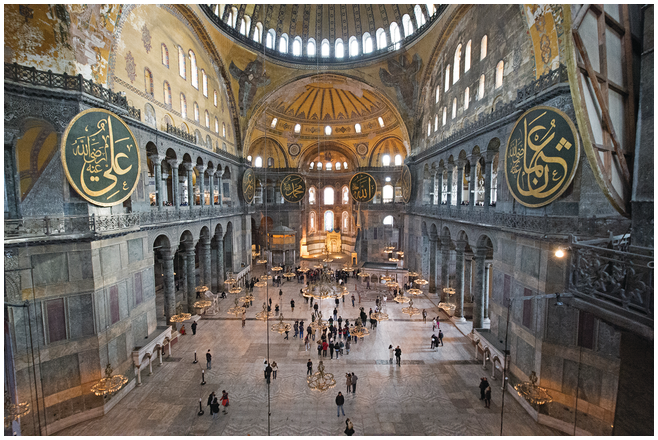 A top-down,wide-angle photograph of Hagia Sophia's interior. It shows several people on the large field of grey marble pavement. The multi-colored marble walls are visible, as is a portion of the ring of windows around the dome.