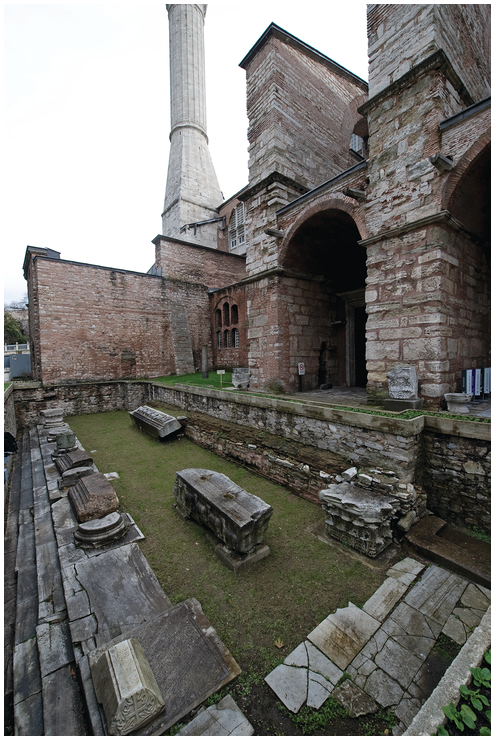 A top-down, wide-angle view of the archaeological remains of the portico of the Theodosian Hagia Sophia. The site is below the current ground level with 3 tiers of mable steps, some paving slabs, portions of column bases, and other fragments.