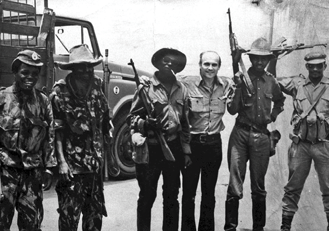 Six men stand side by side, including journalist Ryszard Kapuściński in civilian clothes, surrounded by armed soldiers in uniform carrying long guns. A military truck stands behind them.
