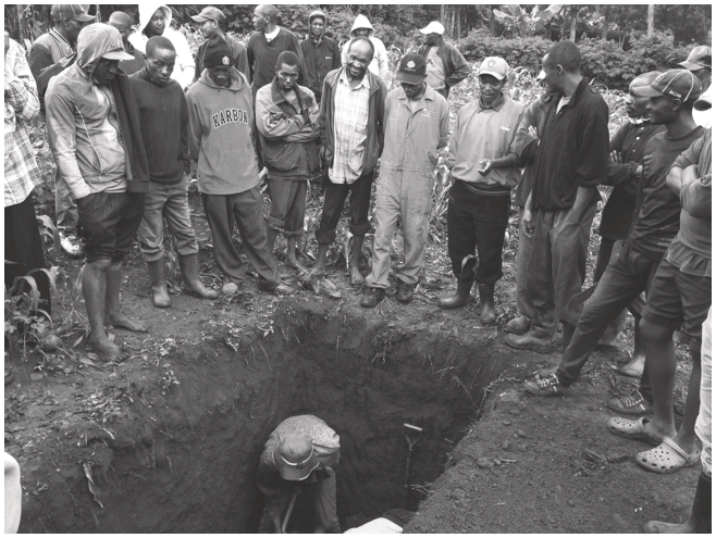 A group of men standing in a circle around a grave being dug by two other men. See long description.