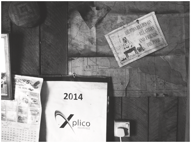 A close-up of the wall of a house, its wooden panels covered by a map and a calendar. On top of the map, there is a sketch of a man squatting on a stool and crying. Next to him, the text reads, ‘He who relies on relatives and friends die poor’. See long description.