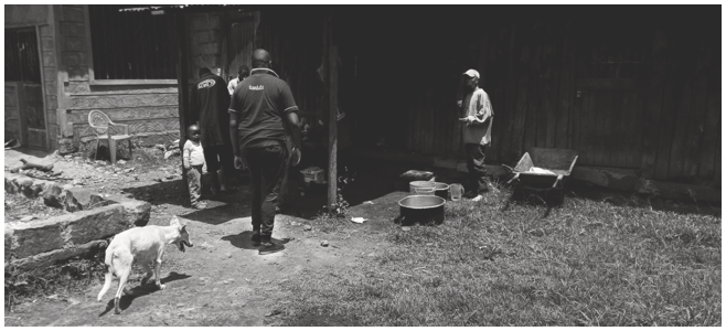 A photo of a man as he approaches a homestead. To his left, a small girl and a dog are visible, with other figures in the shadows. To his right, another man stands next to a wheelbarrow and cooking pots on the grass-covered ground. See long description.