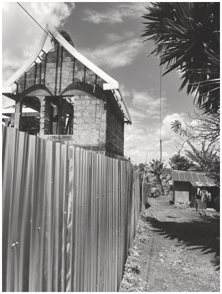 A photo of a tall, unfinished, two-storey house, as seen from behind a corrugated iron fence, made of stone overshadows, belonging to a visibly poorer family. See long description.
