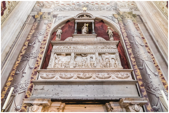 A marble wall tomb viewed from below. The rectangular sarcophagus has three carved reliefs and ornamental floral patterns. Three statues, including a bishop under an architectural canopy, sit atop it. Frescoed columns frame the wall tomb.