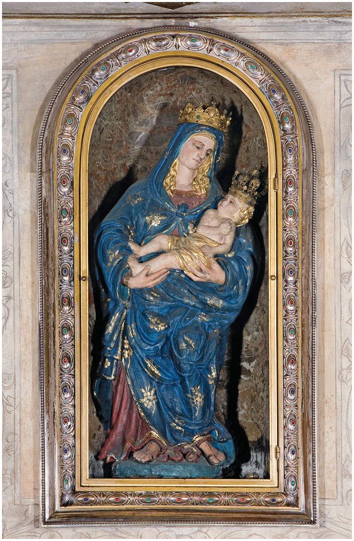 A painted sculpture of the Virgin Mary, standing and holding the Christ Child in her arms, in an arched wall niche. She wears blue robes with gold stars. Both figures have blond hair and gold crowns on their heads.