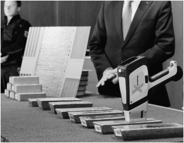 A series of 8 gold bars laid out on a table, with men in suits standing behind it. An Olympus handheld analyzer to test the purity of the bars, is placed on top of one of the bars.