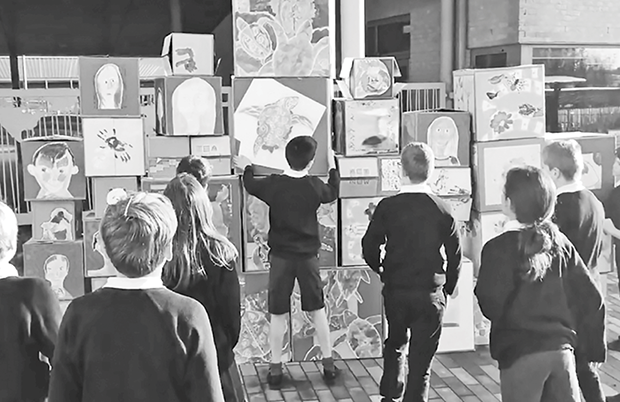 A group of primary school children in school uniform are gathered around a wall of cardboard boxes decorated with children’s art. They look on while a boy arranges a painting in the centre of the wall.
