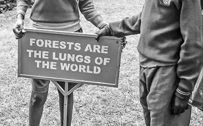 Two young children in school uniforms hold a metal sign. The sign’s bold capital letters state that forests are the lungs of the world. The sign has a stake to hold it in the ground.