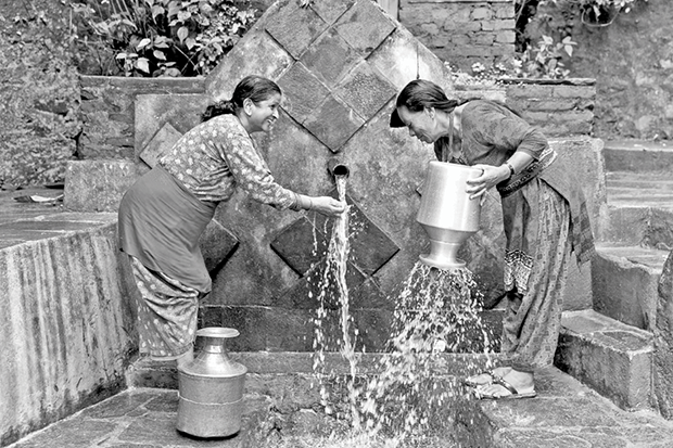 Two women engaged in washing and cleaning metal containers using the directed stream of water from a stone fountain. They are surrounded by stone steps and walls, with vegetation growing in the background.