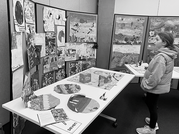 A young girl looks at a display of children’s art arranged on tables and screens. The art depicts landscapes and animals and uses a range of materials to make the pictures three dimensional.