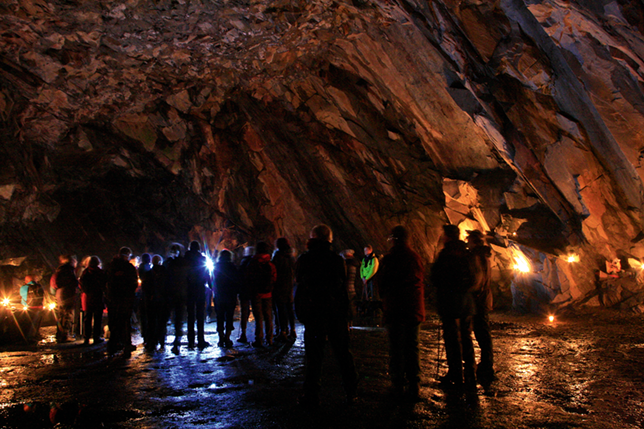 Silhouettes of a group of men and women clustered together within a large cave. The walls of the cave are orange/brown and illuminated by lamps at the side. The floor of the cave is wet.