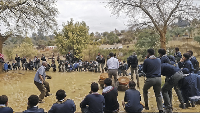 A group of students on school uniform in a squatting position, but some standing in a dancing posture forming a circle. Two boys and the teacher are in the middle. The teacher has one foot raised, in a dancing posture.