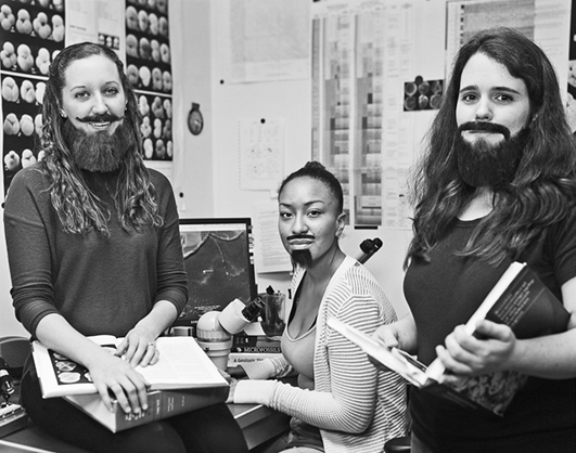 Three young women, wearing fake beards, are posing by a desk with a microscope. They are working on microfossils. Two of them have opened books in their hands. The third one, in the middle of the picture, is sitting in front of the microscope.