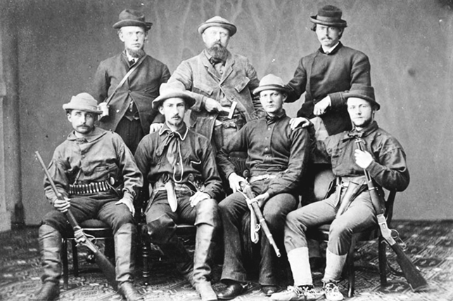 Seven men are posing for a group portrait. In the front, four are seating on chairs with riffles in their hands, except for one. The other three are standing in the back. Othniel C. Marsh in the middle is holding a geologist’s hammer by his waist.