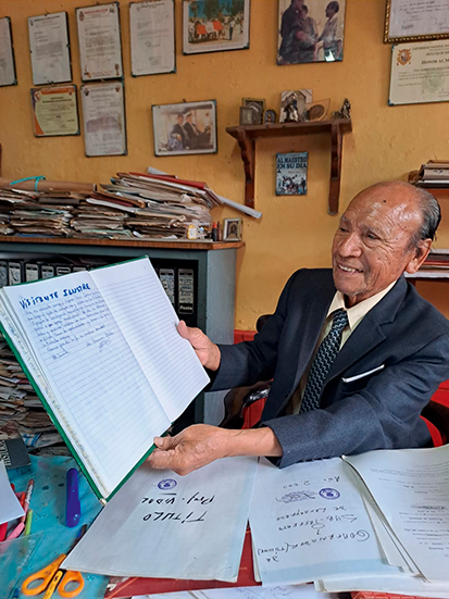 A portrait of Professor Vidal Cobeñas, seated in his office, surrounded by books and research materials. He smiles and holds up an open book.