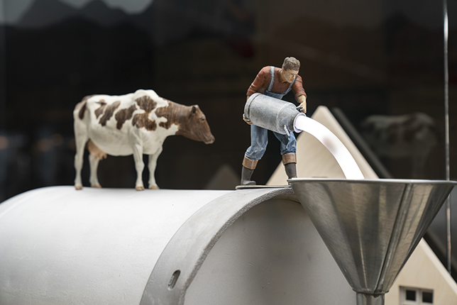 A miniature sculpture showing a man pouring milk from a milk container into an industrial cylinder. A miniature cow stands by.