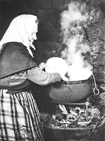 A woman in front of a stone stow heating and stirring milk in a pot.