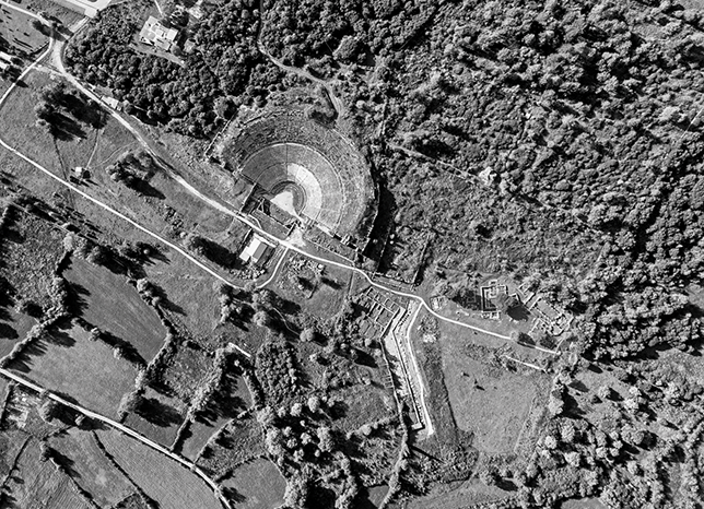 An aerial photograph of ruins surrounded by trees. A road runs across the site. There is a theatre and some buildings. See long description.